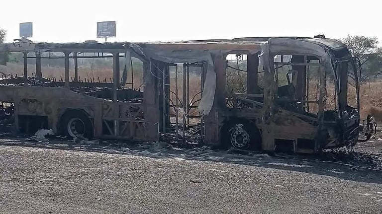 Two charred, burnt-out buses on a barren ground with buildings visible in the background