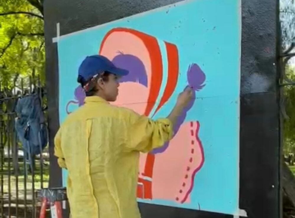 Artist in yellow shirt and blue cap painting colorful abstract mural on large turquoise panel outdoors