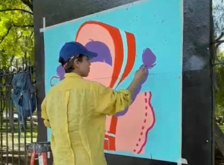 Artist in yellow shirt and blue cap painting colorful abstract mural on large turquoise panel outdoors