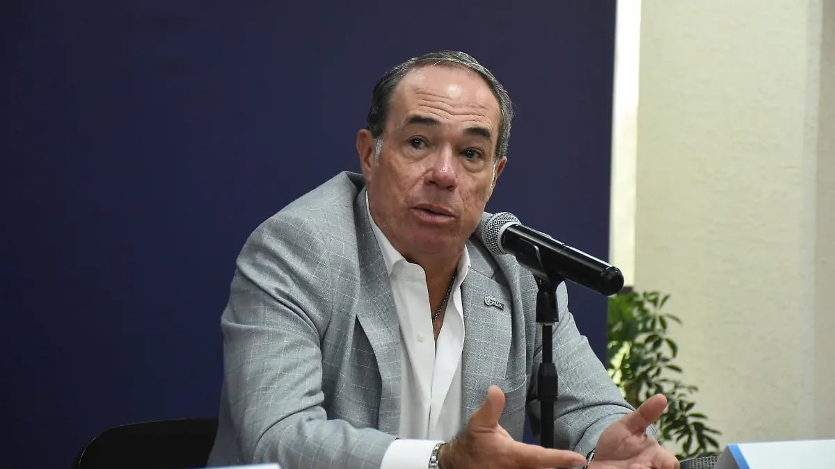 Man in gray blazer speaking into microphone at a table during a presentation or conference