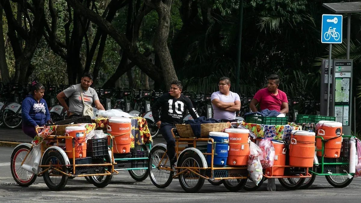 A group of people sitting on colorful orange cargo bikes loaded with beverages and supplies under trees on a city street