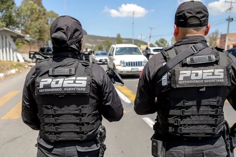 Two police officers wearing tactical gear and helmets stand on a road with parked vehicles in the background