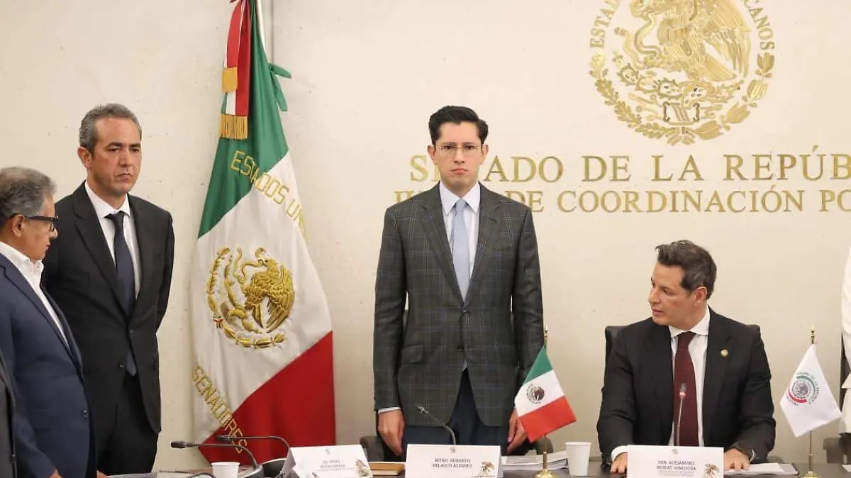 Four men in business suits at an official government meeting with Mexican flags and seals visible in background