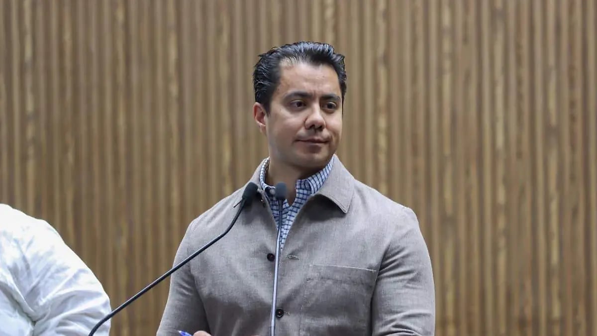 Man in gray jacket speaking at microphone against beige textured wall