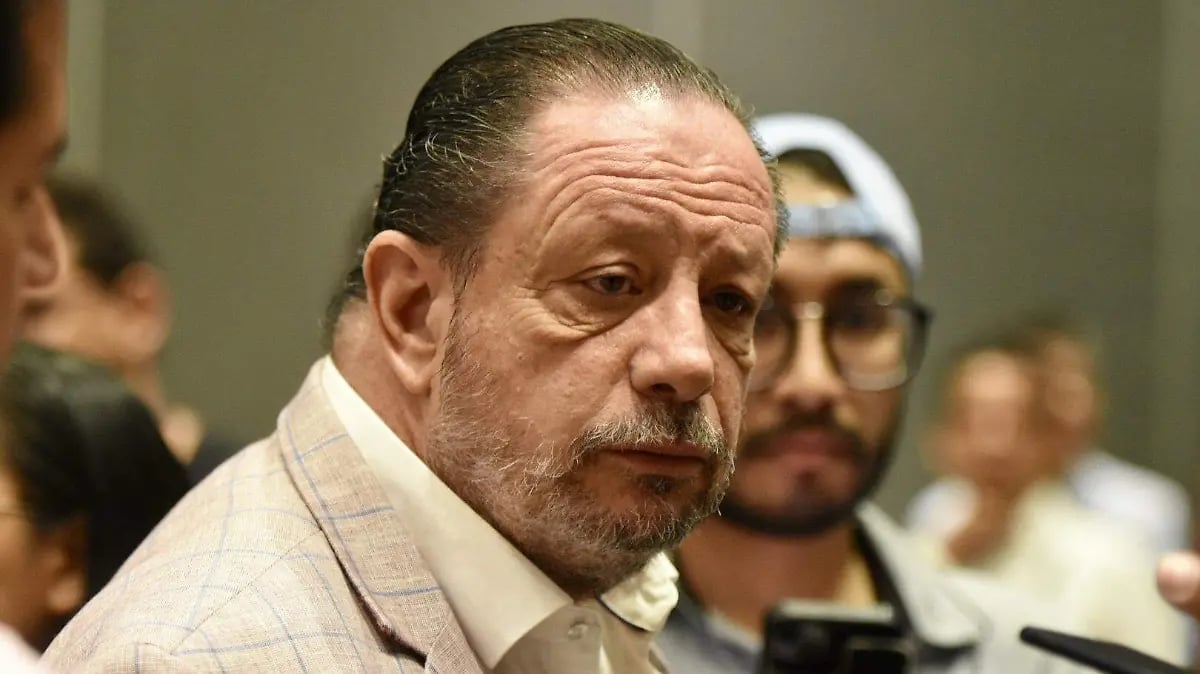 Middle-aged man with gray hair and mustache wearing a light tan jacket in what appears to be a courtroom or official setting